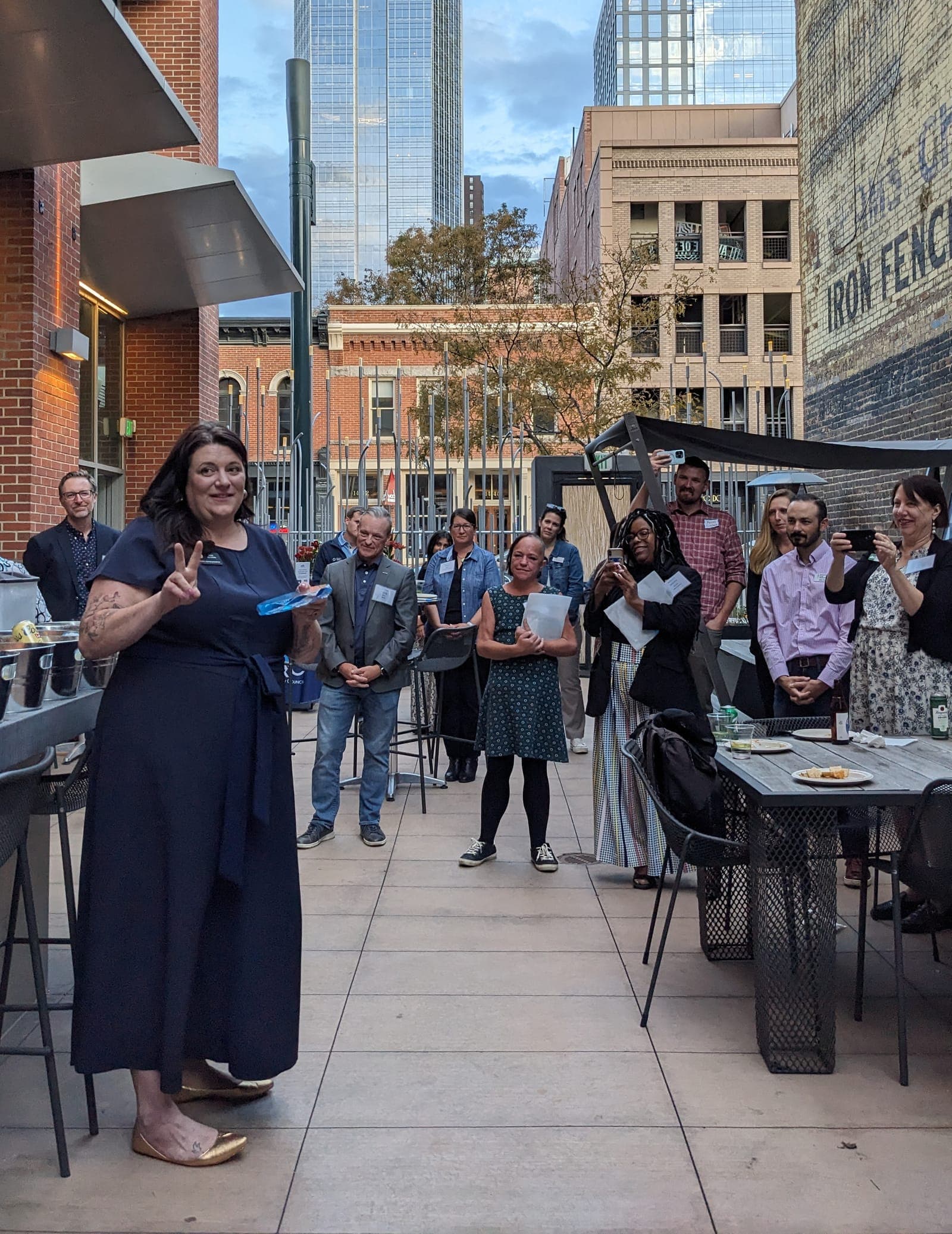 A guest holds up an award and a peace sign at an evening reception on a downtown Denver patio as guests look on