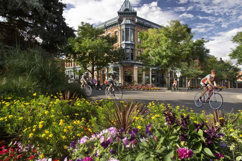 Cyclists ride past a Victorian brick corner building in Old Town Fort Collins, with bright flowerbeds in the foreground