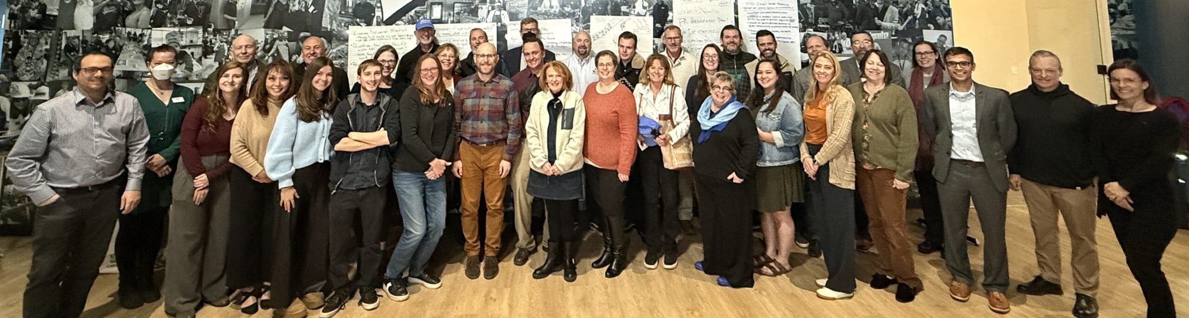 The RAQC Board and staff in 2025, about 40 people standing together in front of a black-and-white photo mural and meeting notes.