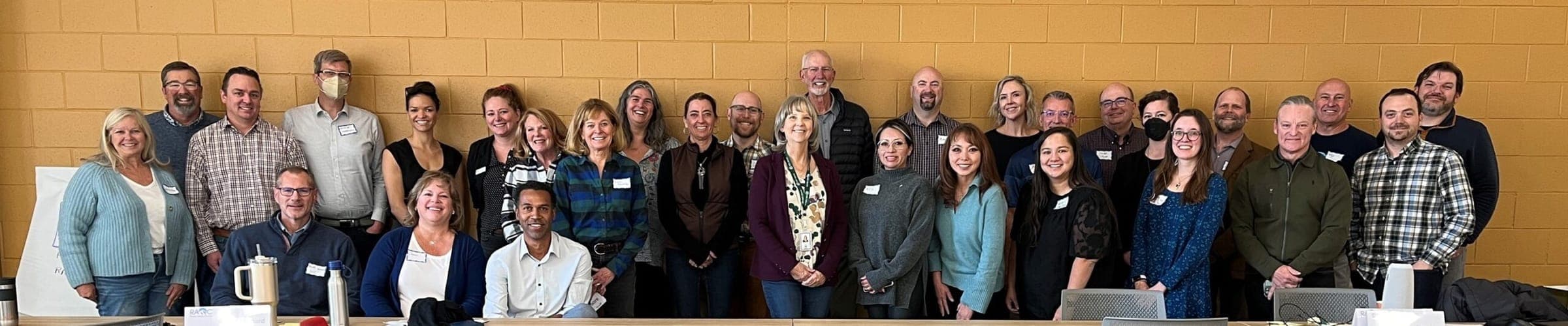 The RAQC Board and staff in November 2024, about 35 people standing and seated together in front of a yellow brick wall.